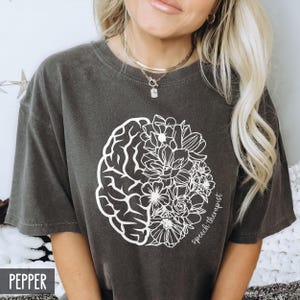 May include: A dark gray t-shirt with a white outline of a brain and flowers. The text "speech therapist" is written on the shirt.