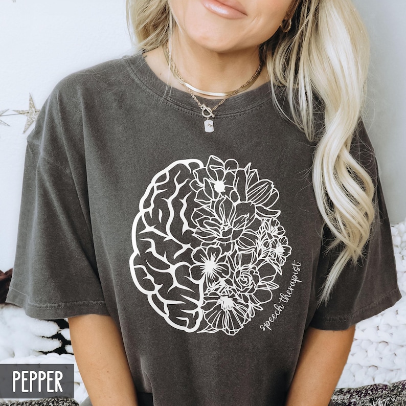 May include: A dark gray t-shirt with a white outline of a brain and flowers. The text "speech therapist" is written on the shirt.