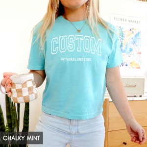 May include: A turquoise blue cropped t-shirt with the word "CUSTOM" in white outlined letters. The shirt has an optional second line that says "OPTIONAL 2ND LINE".