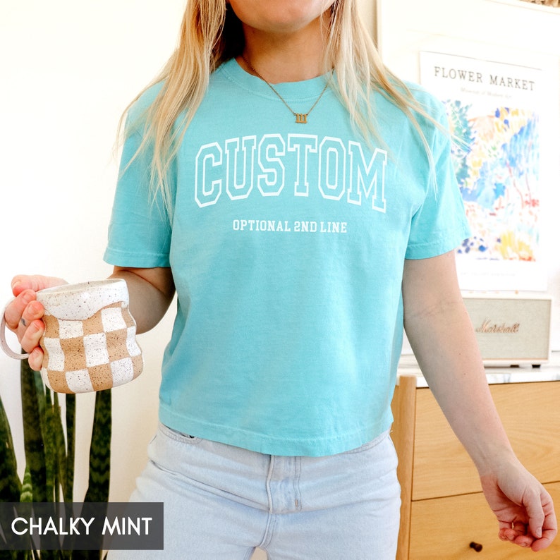 May include: A turquoise blue cropped t-shirt with the word "CUSTOM" in white outlined letters. The shirt has an optional second line that says "OPTIONAL 2ND LINE".