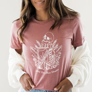May include: A dusty rose t-shirt with a white graphic of a skeleton holding a book. The graphic is surrounded by floral elements. The text "Just One More Chapter" is printed below the graphic.