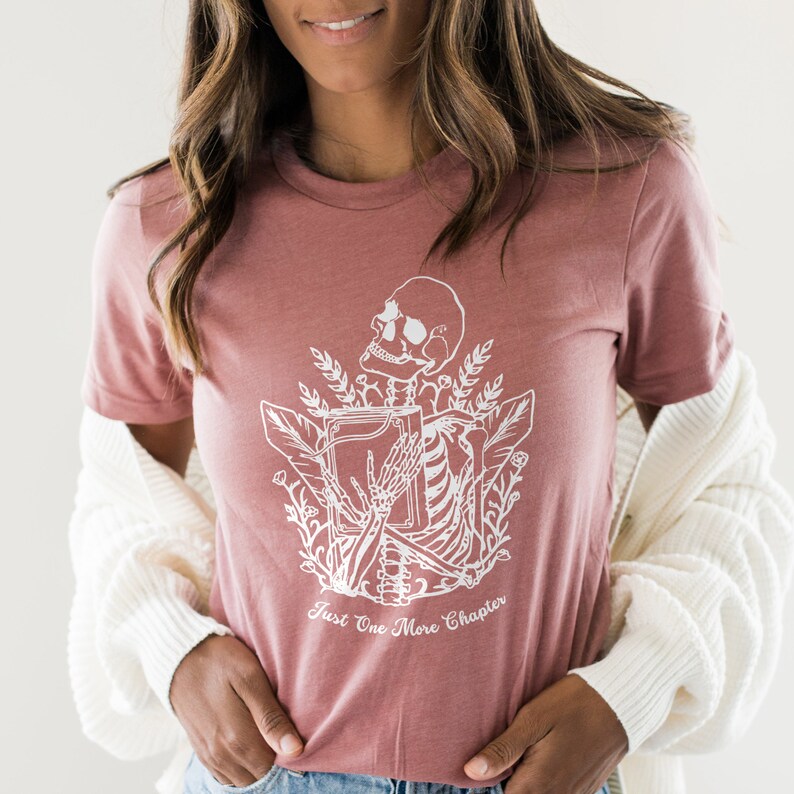 May include: A dusty rose t-shirt with a white graphic of a skeleton holding a book. The graphic is surrounded by floral elements. The text "Just One More Chapter" is printed below the graphic.
