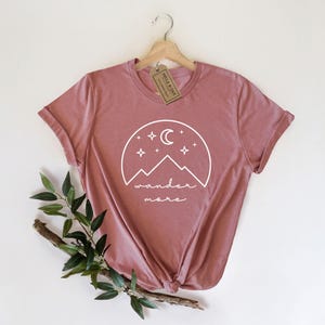 A mauve t-shirt hanging on a wooden hanger displays a white mountain and moon graphic with the text wander more.