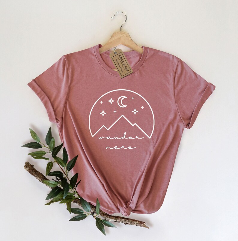 A mauve t-shirt hanging on a wooden hanger displays a white mountain and moon graphic with the text wander more.
