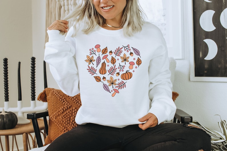 May include: White sweatshirt with a heart-shaped design featuring autumn leaves, acorns, pumpkins, and mushrooms in shades of orange, pink, and brown.