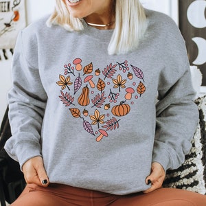 May include: A grey sweatshirt with a heart-shaped design featuring autumnal elements like leaves, pumpkins, acorns, and mushrooms in shades of orange, brown, pink, and yellow.
