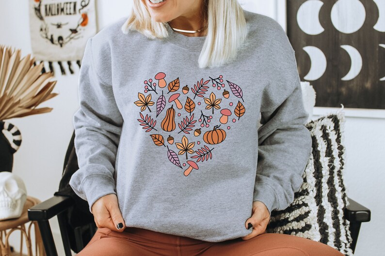 May include: A grey sweatshirt with a heart-shaped design featuring autumnal elements like leaves, pumpkins, acorns, and mushrooms in shades of orange, brown, pink, and yellow.