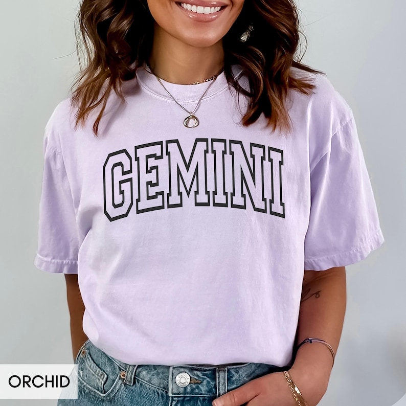 May include: Orchid-colored t-shirt with the word "GEMINI" in a black, block-letter font. The shirt is a light purple color and has a relaxed fit. The word "ORCHID" is printed at the bottom of the image.