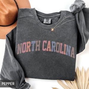 Puede incluir: Una sudadera gris oscuro con el texto "NORTH CAROLINA" en un degradado de rosa, naranja y azul. La sudadera tiene cuello redondo y mangas largas. Un collar dorado con un colgante est&aacute; sobre la camisa.