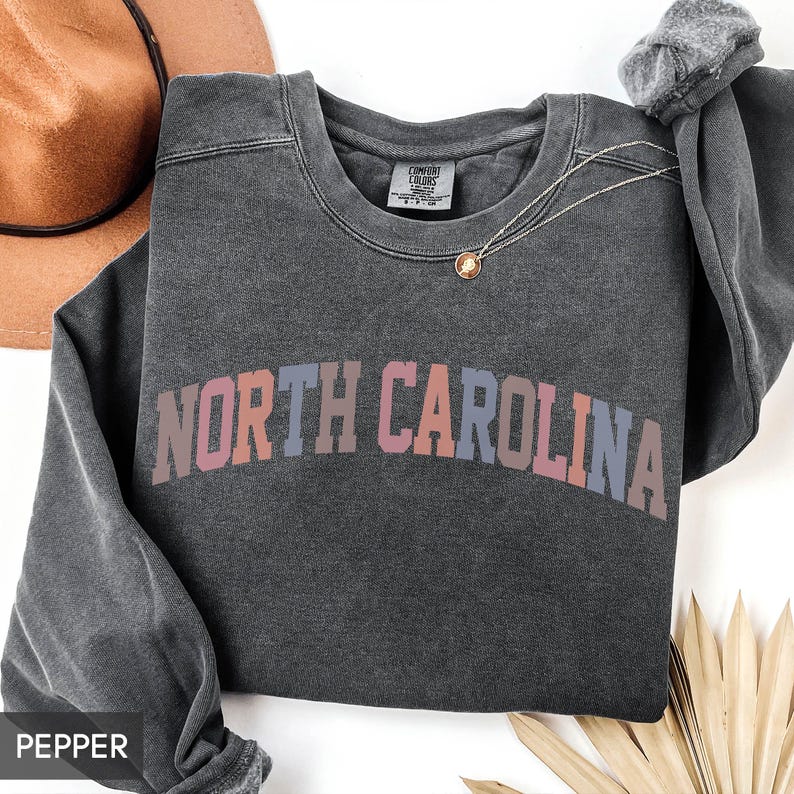 Puede incluir: Una sudadera gris oscuro con el texto "NORTH CAROLINA" en un degradado de rosa, naranja y azul. La sudadera tiene cuello redondo y mangas largas. Un collar dorado con un colgante est&aacute; sobre la camisa.