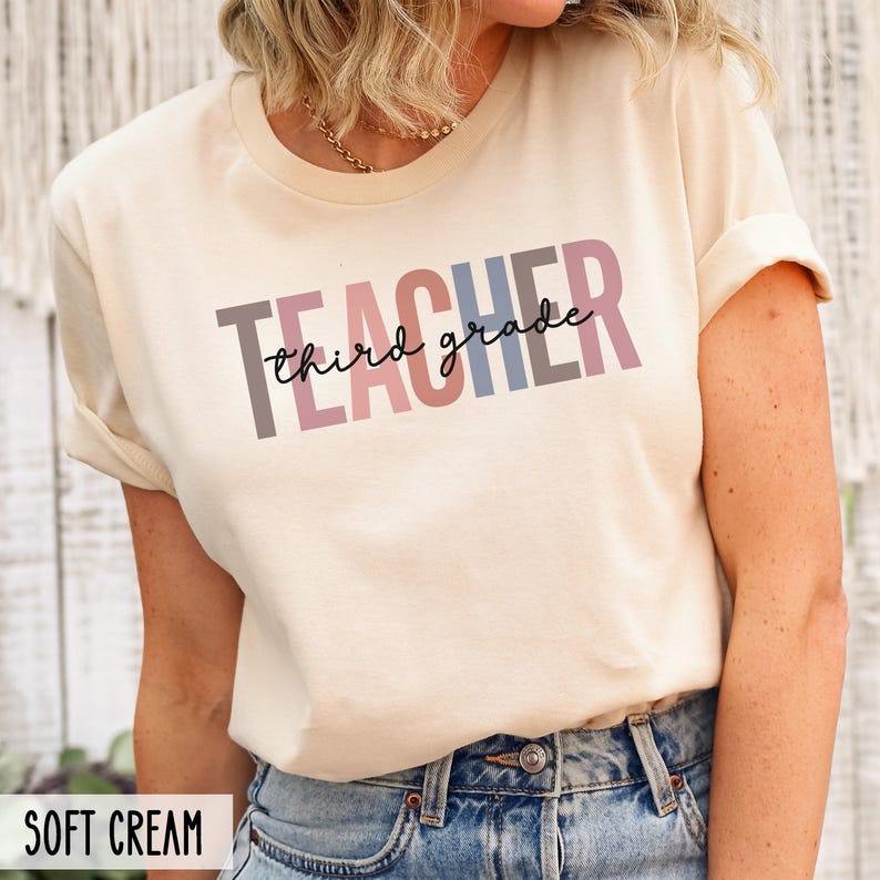 A woman wearing a soft cream t-shirt with multicolored block letters spelling teacher and cursive text reading third grade across the chest.