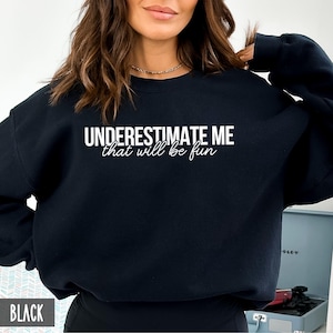 May include: Black sweatshirt with the text "UNDERESTIMATE ME that will be fun" in white.