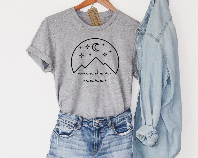 Pode incluir: Camiseta cinza de manga curta com um gr&aacute;fico preto de montanhas, uma lua crescente e estrelas. O texto "wander more" est&aacute; escrito abaixo do gr&aacute;fico.