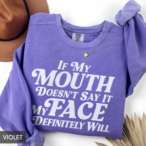 Puede incluir: Una sudadera violeta con el texto blanco: "If My Mouth Doesn't Say It My Face Definitely Will". La sudadera de cuello redondo tiene mangas largas. Un collar dorado con un colgante est&aacute; drapeado en la parte delantera.