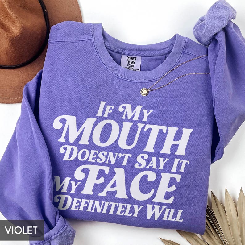 Puede incluir: Una sudadera violeta con el texto blanco: "If My Mouth Doesn't Say It My Face Definitely Will". La sudadera de cuello redondo tiene mangas largas. Un collar dorado con un colgante est&aacute; drapeado en la parte delantera.