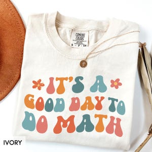 May include: Ivory-colored t-shirt with the phrase "IT'S A GOOD DAY TO DO MATH" in colorful, retro-style lettering. The shirt has a crew neck and short sleeves. A gold necklace with a pendant is draped across the shirt.