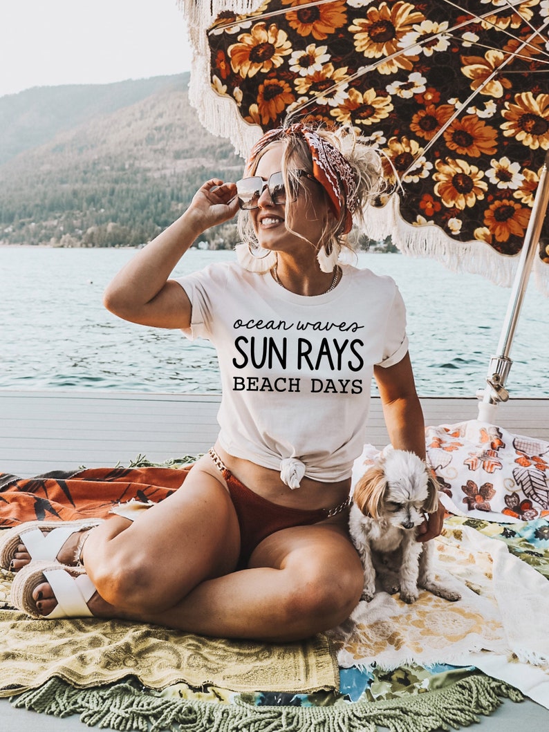 May include: A woman wearing a white t-shirt with the text "ocean waves SUN RAYS BEACH DAYS" sits on a blanket with a small white dog. She is wearing sunglasses and a red bandana in her hair. The woman is sitting under a floral umbrella.