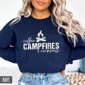 A person wears a navy crewneck sweatshirt featuring white text that reads coffee CAMPFIRES & cocktails with a campfire graphic.