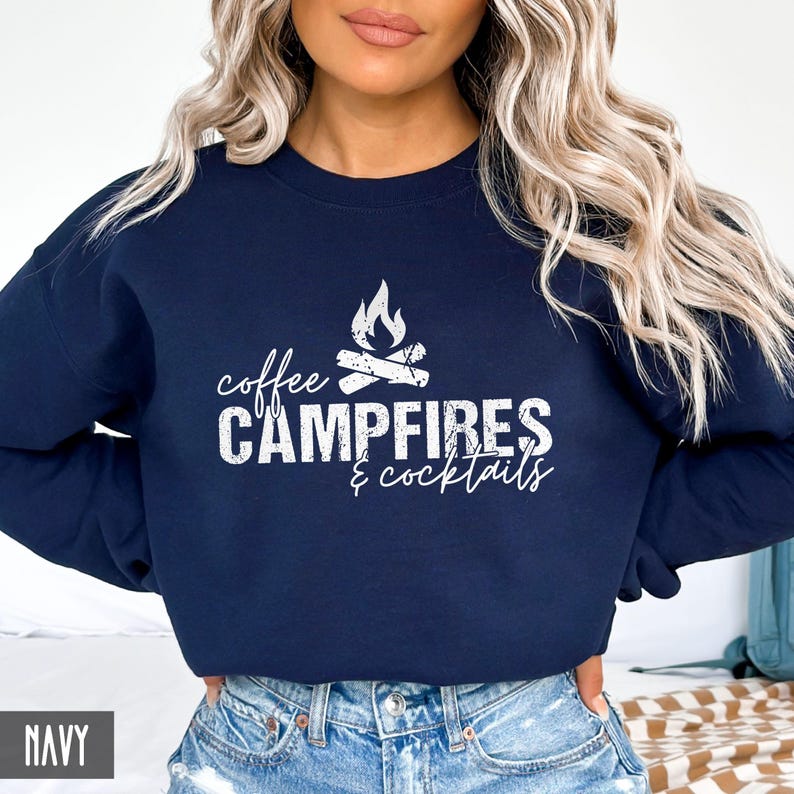 A person wears a navy crewneck sweatshirt featuring white text that reads coffee CAMPFIRES & cocktails with a campfire graphic.