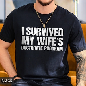 May include: Black t-shirt with the white text "I SURVIVED MY WIFE'S DOCTORATE PROGRAM". The shirt is a crew neck and is being worn by a person. A gold chain necklace is visible.