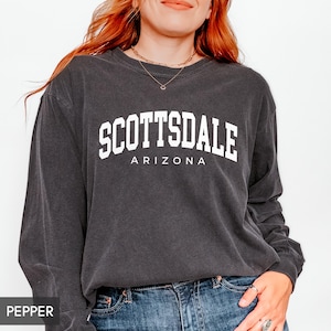 May include: A dark gray long-sleeve sweatshirt with the word "SCOTTSDALE" in large white block letters above "ARIZONA" in smaller white letters. The sweatshirt is worn with blue jeans.