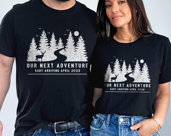 Puede incluir: Una camiseta negra de manga corta con un gráfico blanco de un bosque, un camino sinuoso, un ciervo y una luna. El texto "OUR NEXT ADVENTURE" y "BABY ARRIVING APRIL 2025" aparece en letras blancas debajo de la ilustración de la naturaleza.