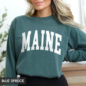 May include: A blue spruce sweatshirt with the word "MAINE" in large white block letters across the chest. The sweatshirt is a crew neck style and has long sleeves. The text "BLUE SPRUCE" is at the bottom of the image.