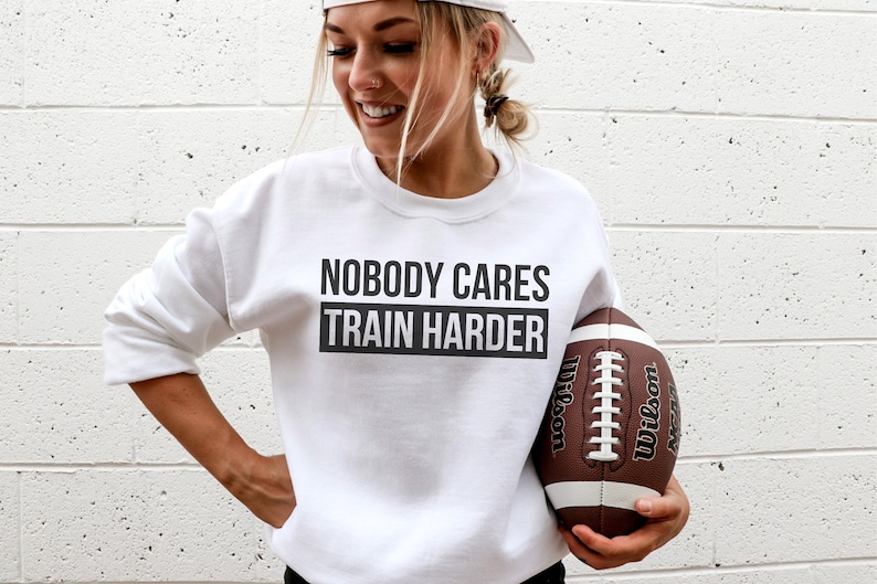 Pu&ograve; includere: Felpa bianca con il testo "Nobody Cares Train Harder" in lettere nere. Una persona tiene un pallone da football marrone e bianco.