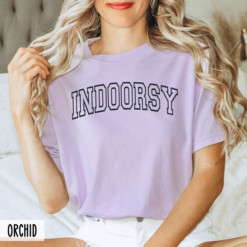 May include: A lavender t-shirt with the word "INDOORSY" printed in black block letters.