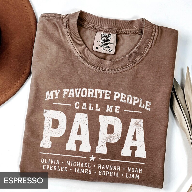 A brown t-shirt with a humorous text printed on it, which reads "MY FAVORITE PEOPLE CALL ME PAPA" and lists the names of people who are considered "Papa" in the text.
