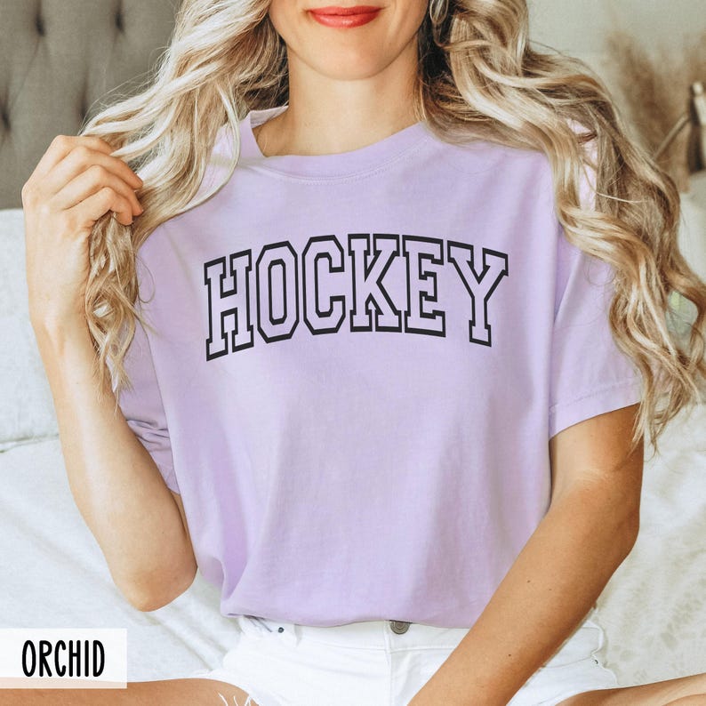 A woman wearing an orchid purple t-shirt with black block letters that spell HOCKEY.