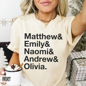 K&ouml;nnte beinhalten: Ein cremefarbenes T-Shirt mit schwarzem Text, der "Matthew & Emily & Naomi & Andrew & Olivia." lautet.