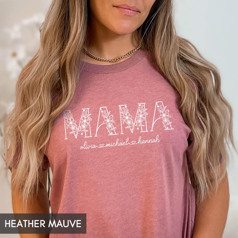 May include: Heather mauve t-shirt with the word "MAMA" in white floral letters. Below the word "MAMA" are the names "olivia", "michael", and "hannah" with heart symbols. The shirt is a soft, heathered fabric.