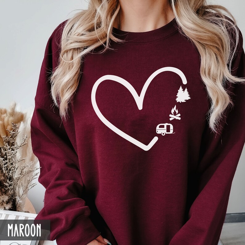 May include: A maroon sweatshirt with a white heart design. Inside the heart are illustrations of a camper, a campfire, and a pine tree.