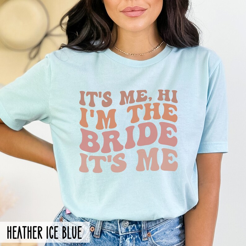 May include: A light blue t-shirt with the text "It's me, hi I'm the bride it's me" in a retro font with pink and orange color gradients. The text is centered on the shirt.