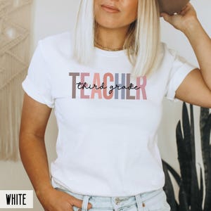 A woman modeling a white t-shirt with multicolored block letters spelling teacher overlaid with black cursive text reading third grade.