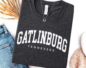 May include: A dark grey heather t-shirt featuring the white text GATLINBURG TENNESSEE in a collegiate font style. The shirt has a crew neckline and is displayed with a silver sunburst pendant necklace resting on the collar area of the garment.