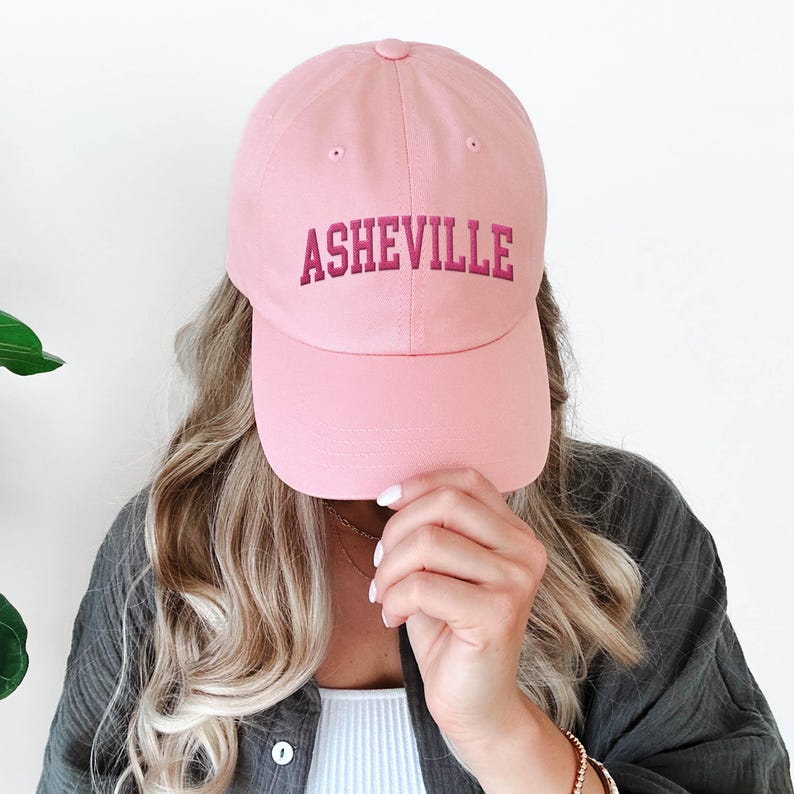 Puede incluir: Gorra de b&eacute;isbol rosa claro con la palabra "ASHEVILLE" bordada en rosa oscuro. La gorra tiene una visera curva y est&aacute; hecha de una tela suave. Un estilo cl&aacute;sico, adecuado para ropa informal.