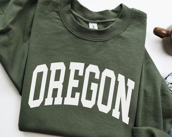 May include: A military green sweatshirt with the word "OREGON" printed in large, white, block letters across the front. The sweatshirt is folded, with a portion of a denim garment visible. The text "MILITARY GREEN" is at the bottom.
