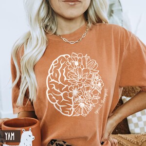 May include: A burnt orange t-shirt with a white graphic of a brain and flowers. The text "speech therapist" is written on the shirt.