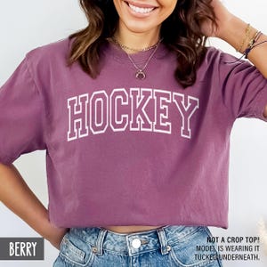 A woman wearing a berry purple t-shirt with white block letters that spell HOCKEY.