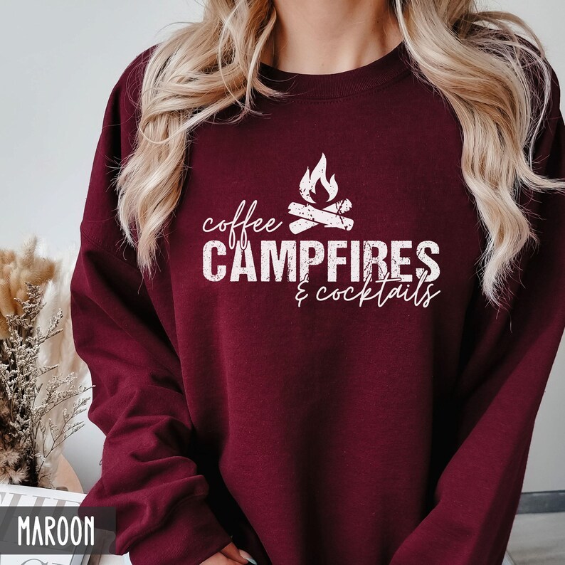 A person wears a maroon crewneck sweatshirt featuring white text that reads coffee CAMPFIRES & cocktails with a campfire graphic.