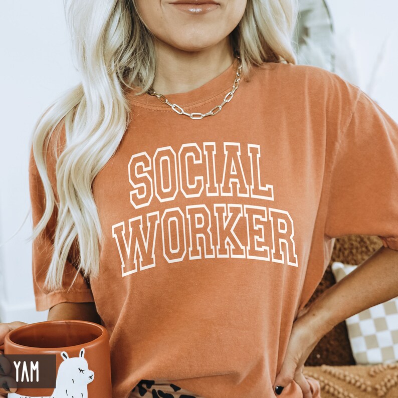 Puede incluir: Una camiseta naranja quemada con el texto "SOCIAL WORKER" impreso en blanco. El texto est&aacute; en un estilo de fuente universitario.