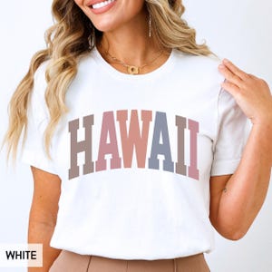 Può includere: Una t-shirt bianca a maniche corte con la parola HAWAII stampata sul petto in un grande carattere arcuato in stile college. Le lettere sono colorate in tonalità alternate e tenui di marrone, rosa polvere e blu ardesia.