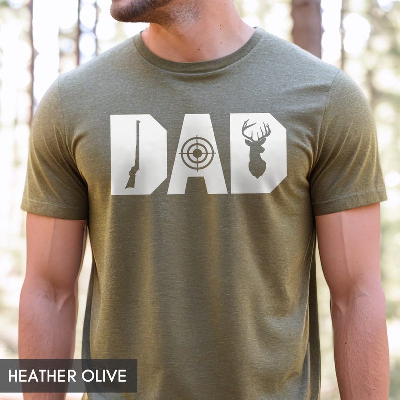 May include: Heather olive green t-shirt with the word "DAD" in white block letters. The "D" features a rifle, the "A" a target, and the last "D" a deer silhouette. The shirt is made of a soft material.
