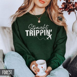 Puede incluir: Una sudadera verde oscuro con el texto "Straight Trippin'" en letras blancas y un gr&aacute;fico de avi&oacute;n blanco. La sudadera la lleva puesta una persona sentada en un sof&aacute;.
