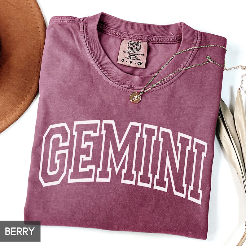 May include: Berry-colored t-shirt with the word "GEMINI" in white block letters. The shirt has a crew neck and a small tag that says "COMFORT COLORS". A gold-colored necklace with a pendant is on the shirt.