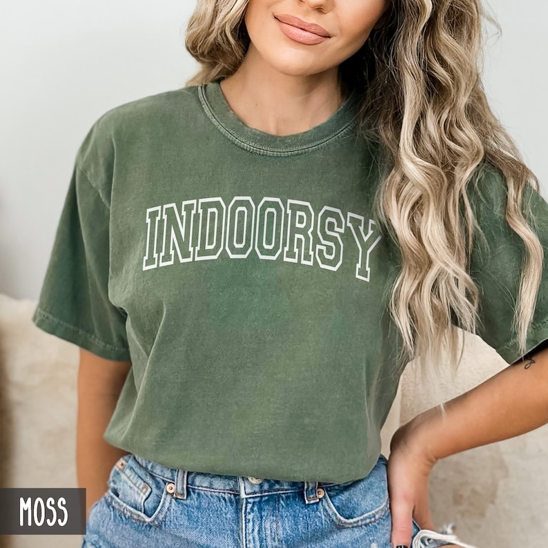 May include: A moss green t-shirt with the word "INDOORSY" printed in white outlined letters.