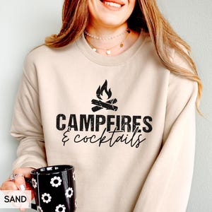May include: A sand-colored sweatshirt with the text "Campfires & Cocktails" in black, along with a graphic of a campfire. A black mug with white flower designs is held in the hand. The word "SAND" is printed in the lower left corner.