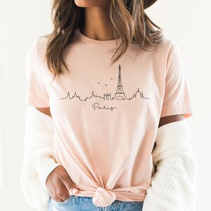Op de afbeelding: Een roze T-shirt met een zwarte lijntekening van de Eiffeltoren en het woord "Paris" in cursief.